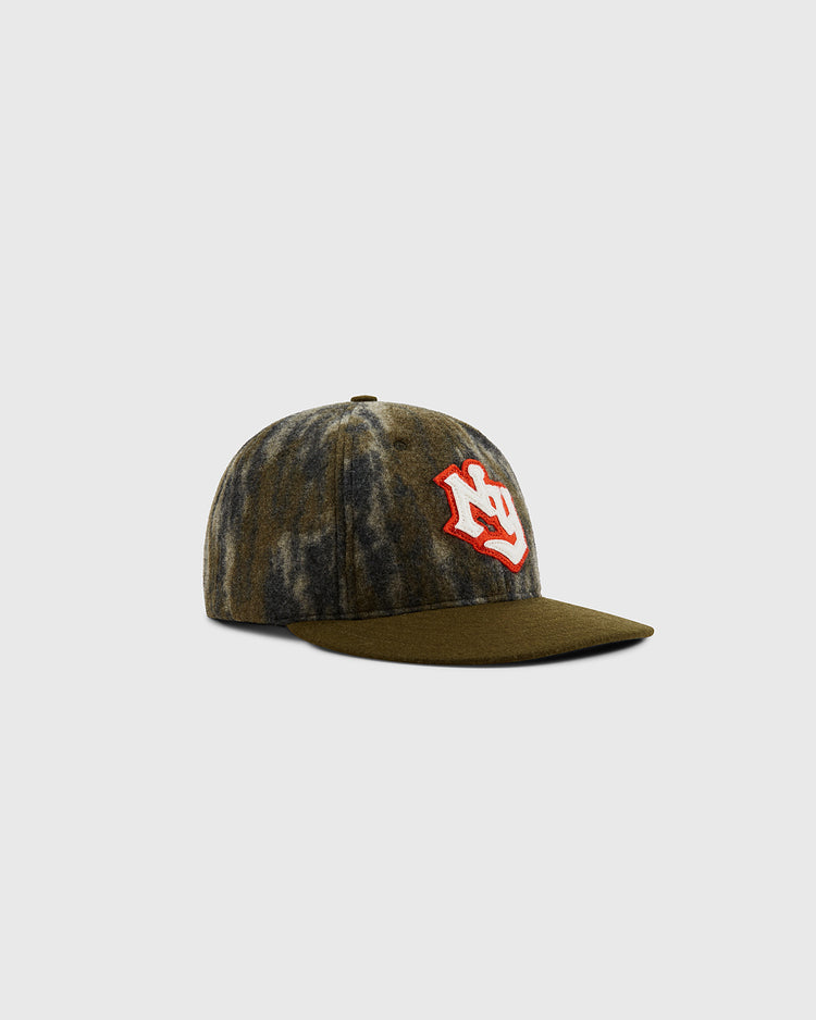 The Mossy Oak x Ebbets Field New York Knights Ballcap by Ebbets Field features a green and gray tie-dye pattern with a curved brim and a bold red and white embroidered logo on the front, displayed on a plain light gray background.