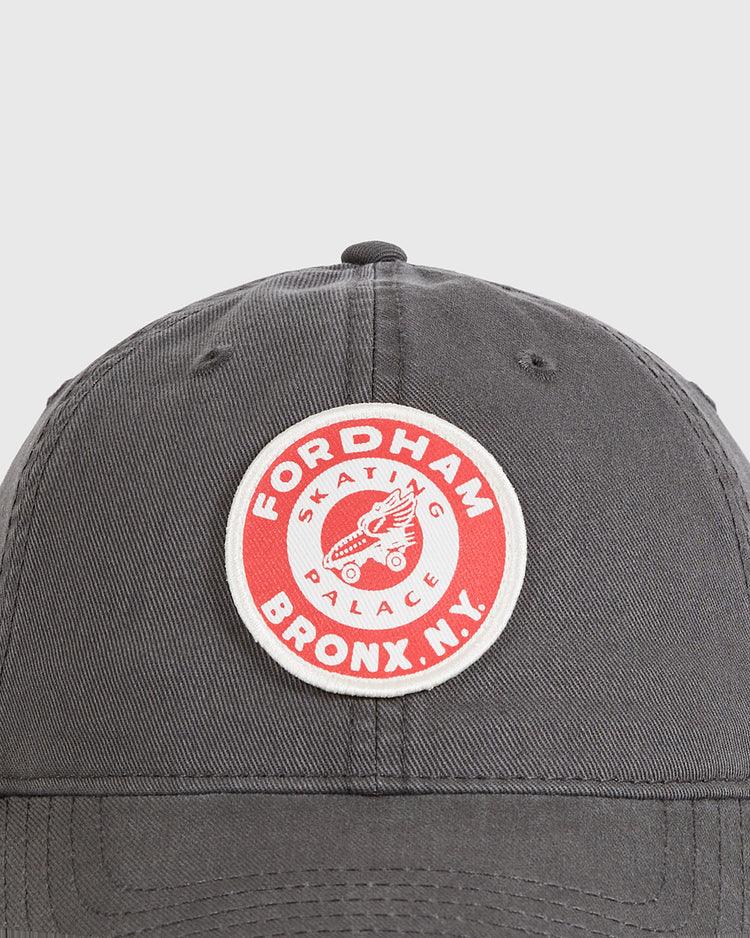 A close-up of the Ebbets Field Fordham Bronx Roller Rink Washed Cotton Ballcap, featuring a dark gray design with a round red and white patch reading 