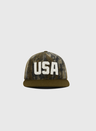 The Ebbets Field Mossy Oak x 1956 USA National Team Ballcap features Bottomland camo, a dark brim, and bold white “USA” lettering on the front, displayed against a plain light grey background.