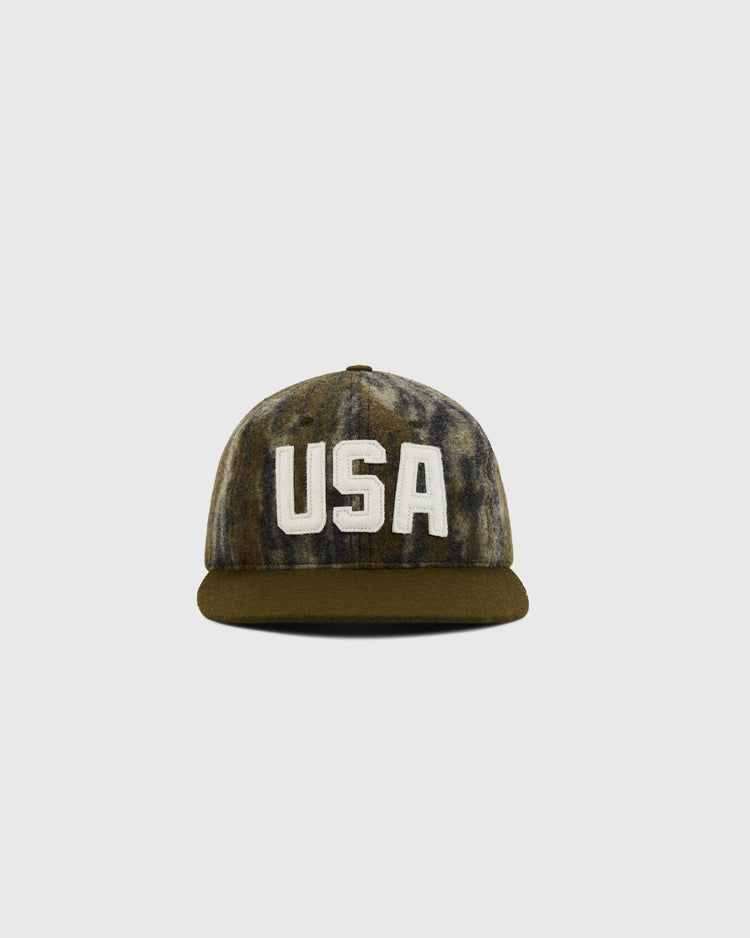 The Ebbets Field Mossy Oak x 1956 USA National Team Ballcap features Bottomland camo, a dark brim, and bold white “USA” lettering on the front, displayed against a plain light grey background.