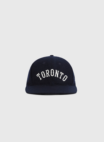 The Ebbets Field Toronto City Series Wool Ballcap features navy blue wool broadcloth and “TORONTO” in white arched block letters on the front, shown against a plain light gray background.
