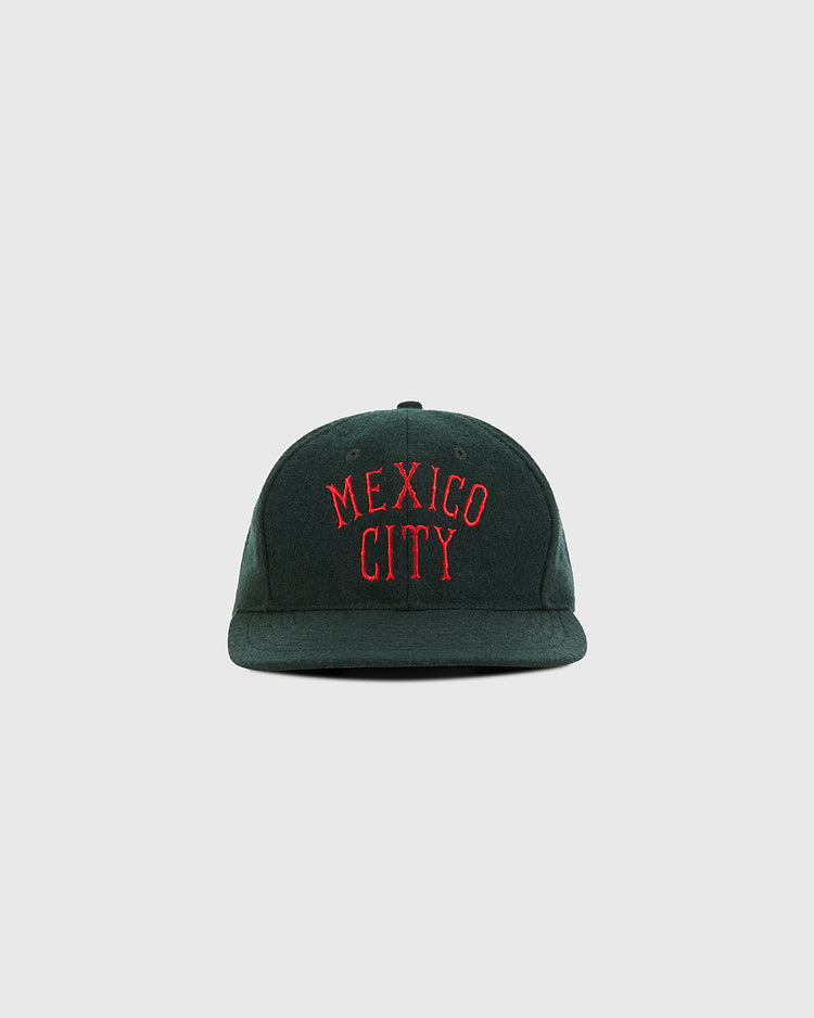 The Ebbets Field Mexico City City Series Wool Ballcap is a dark green hat featuring MEXICO CITY embroidered in red capital letters on the front, shown against a plain light gray background.