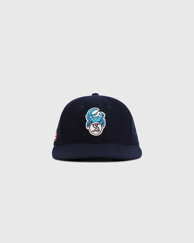 The Santurce Cangrejeros Crab Wool Ballcap by Ebbets Field is a navy blue cap with an embroidered cartoon crab logo in light blue, white, and red, displayed against a plain light gray background.
