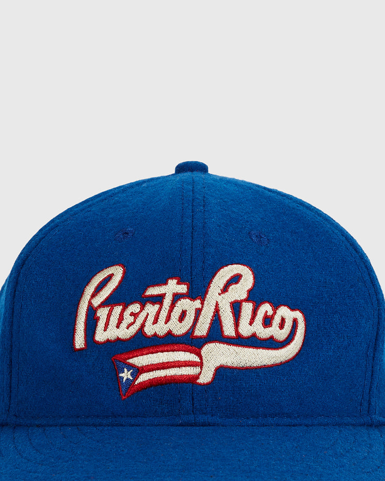 The Ebbets Field Puerto Rico 1972 National Team Wool Ballcap is a blue cap with “Puerto Rico” in white and red script and a stylized flag below, inspired by classic Roberto Clemente-era designs.