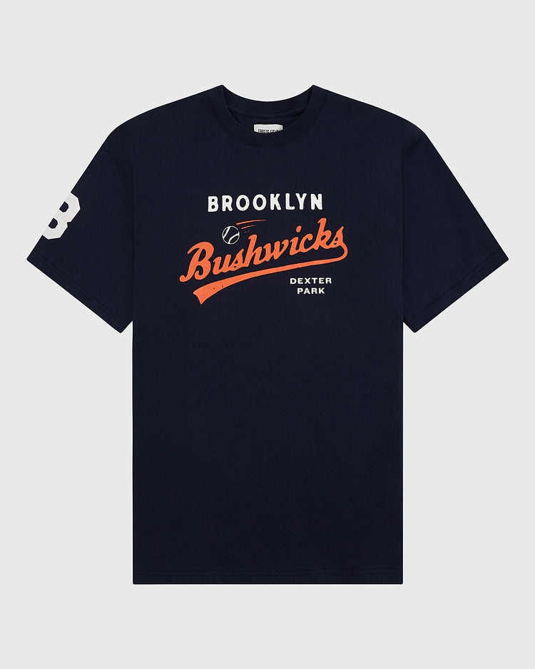 The Ebbets Field Brooklyn Bushwicks Short Sleeve Graphic Tee is a navy vintage-style shirt with “Brooklyn Bushwicks Dexter Park” text, a baseball graphic, and a white number 3 on the left sleeve.