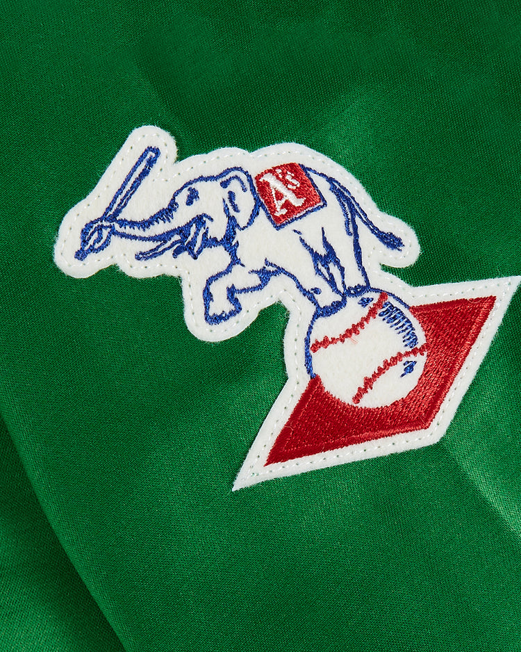 A white patch on green fabric of the EFF Kansas City Athletics 1963 Satin Windbreaker Jacket shows a blue-outlined elephant with a red A’s logo, holding a bat and standing on a baseball atop a diamond—classic 1960s team detail.