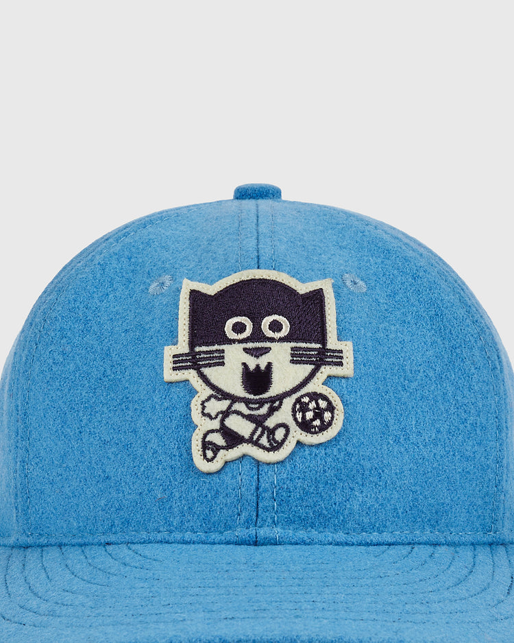 A close-up of the Ebbets Field Chicago Cats Wool Ballcap shows its soft, textured blue fabric and a patch with a smiling cartoon cat in sports clothes kicking a soccer ball on a plain background.