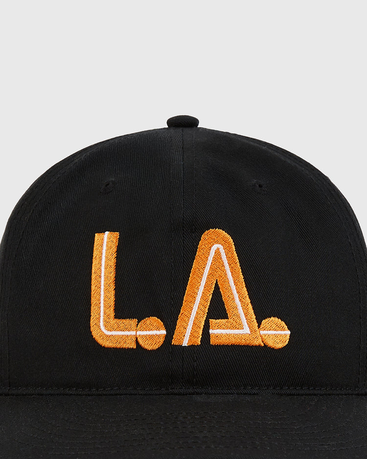 The Ebbets Field Los Angeles Aztecs Cotton Twill Ballcap features “L.A.” in bold orange-yellow embroidery on black cotton twill, nodding to the NASL team. Shown against a light gray background.