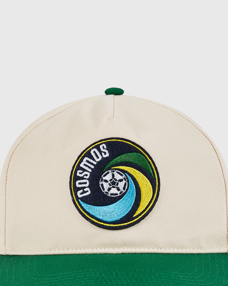 The New York Cosmos A-Frame Cotton Twill Ballcap by Ebbets Field is a beige and green retro snapback featuring an embroidered COSMOS logo with a stylized soccer ball, inspired by the iconic team's classic hat.