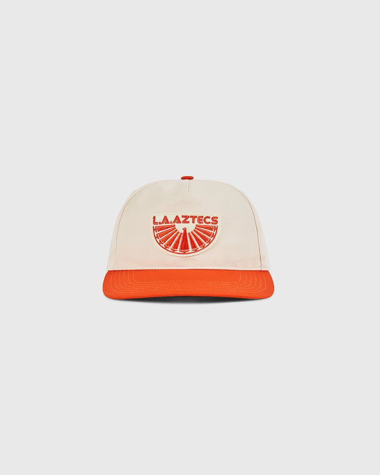 The Ebbets Field Los Angeles Aztecs A-Frame Cotton Twill Ballcap features a beige structured snapback, orange brim, and “L.A. AZTECS” with a sunburst graphic—paying tribute to classic North American Soccer League style.