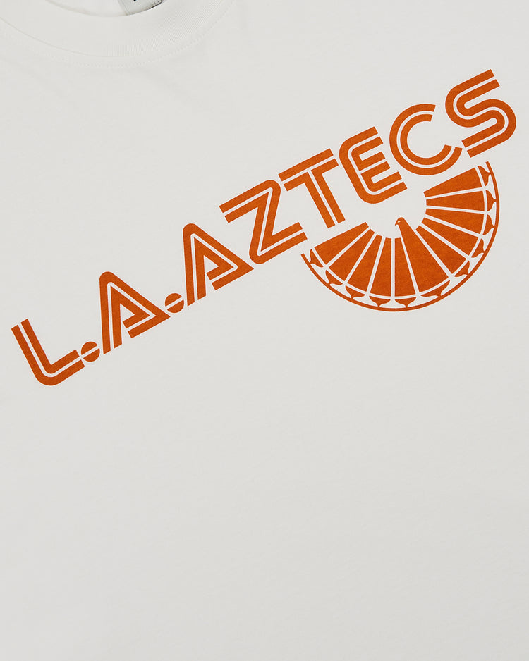 The Los Angeles Aztecs Short Sleeve Graphic T-Shirt by Ebbets Field features bold orange L.A. AZTECS text in a geometric font and a semicircle fan graphic for a vintage soccer look, inspired by the original North American Soccer League team shirt.