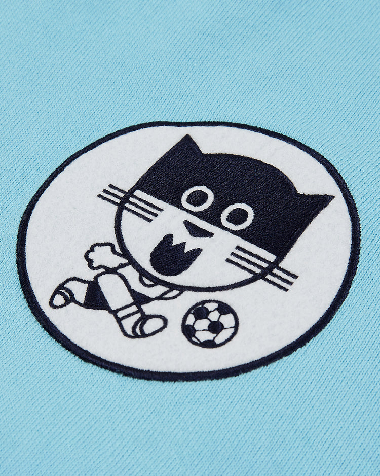 A round patch of the Ebbets Field Chicago Cats Vintage Soccer Goalie Jersey shows a cartoon cat in shorts and shoes, collared long sleeve jersey, open-mouthed with whiskers, playing soccer on a light blue background.