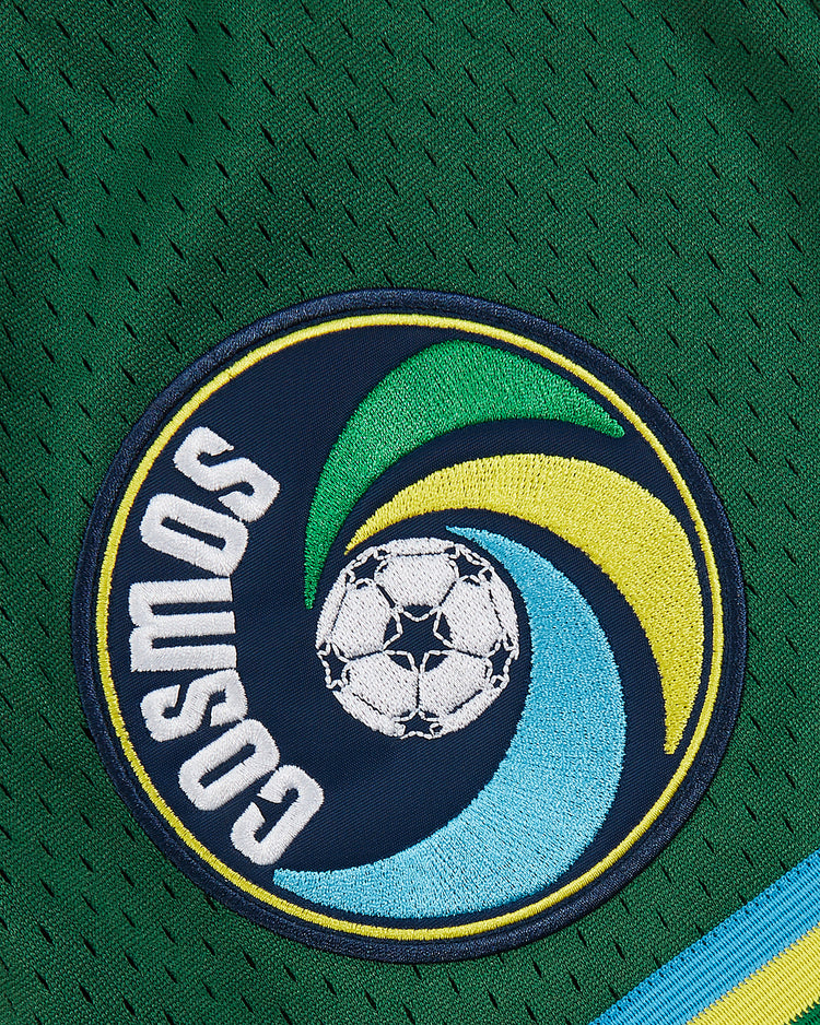 Close-up of green New York Cosmos Mesh Soccer Shorts by Ebbets Field, featuring an embroidered circular patch with COSMOS, a soccer ball, and green, yellow, and blue swirl designs inspired by Pelé's legendary era.