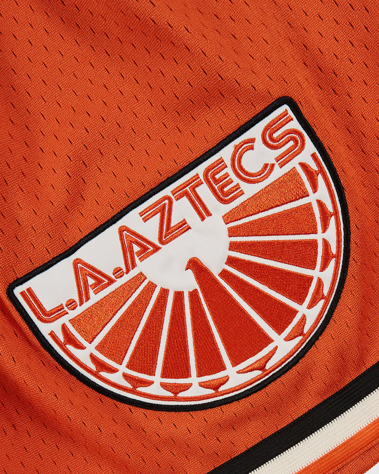 Close-up of orange mesh on Ebbets Field Los Angeles Aztecs Mesh Soccer Shorts, with a retro team patch above a white and red sunburst design.