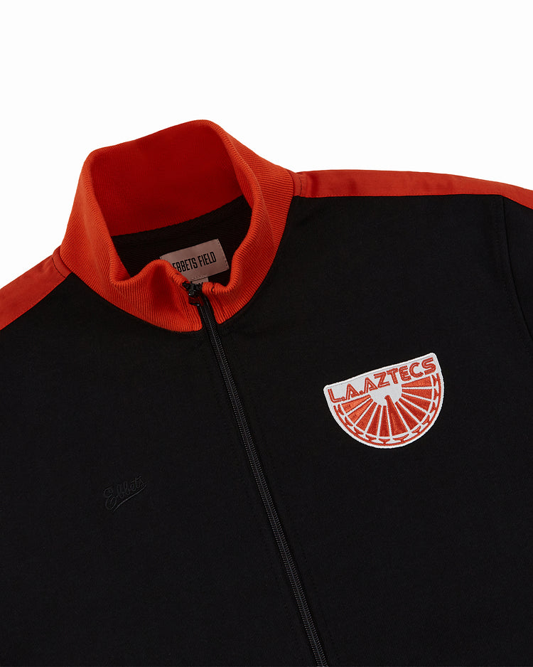 The Ebbets Field Los Angeles Aztecs Soccer Warm Up Jacket is black with a red collar and shoulders, featuring a round LA AZTECS patch with an orange soccer ball on the left chest, inspired by the historic NASL team.
