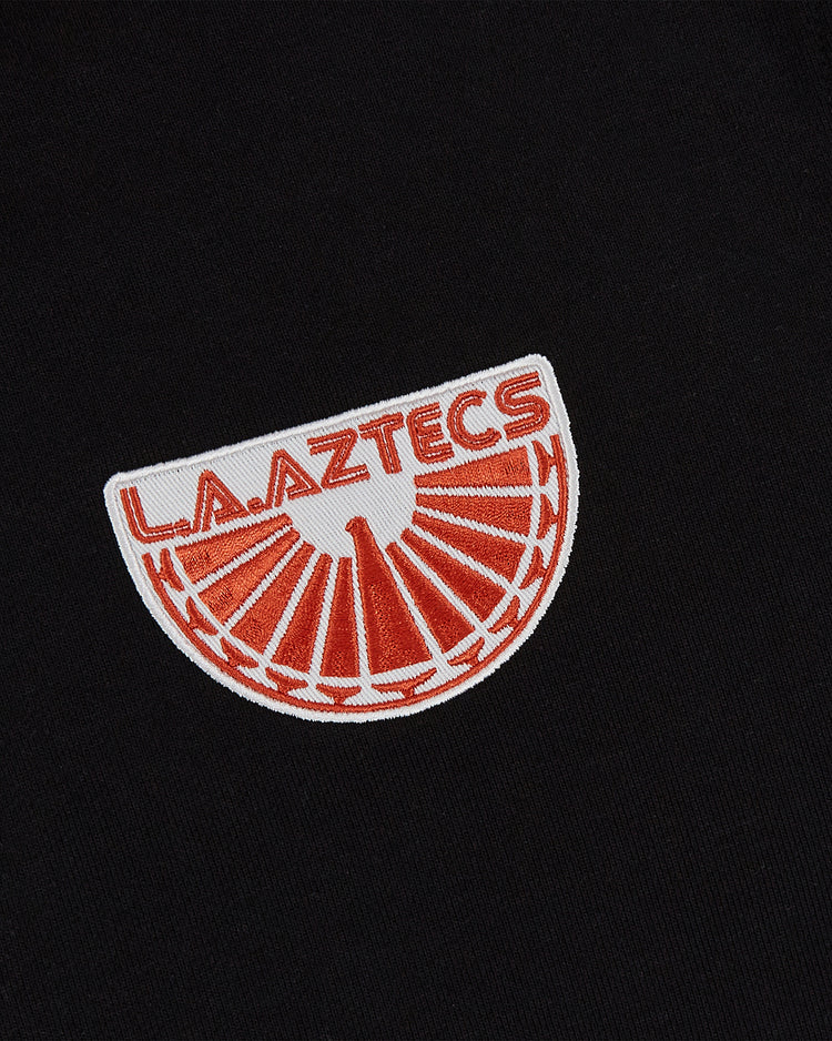 The Ebbets Field Los Angeles Aztecs Soccer Warm Up Jacket features an embroidered L.A. AZTECS patch in red on black, with a red and white sunburst—echoing the classic North American Soccer League design.
