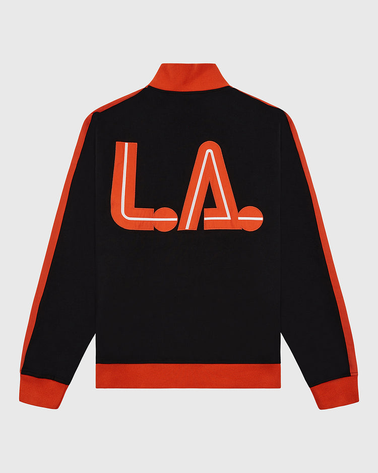 The Ebbets Field Los Angeles Aztecs Soccer Warm Up Jacket is black with red cuffs, collar, and side stripes, and features bold red and white L.A. lettering on the back, inspired by the North American Soccer League team.