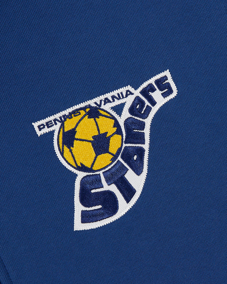 The Ebbets Field Pennsylvania Stoners Soccer Warm Up Jacket is a blue fabric jacket with an embroidered yellow and blue soccer ball and “Pennsylvania Stoners” in white and blue, celebrating ASL champions.