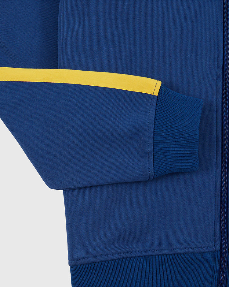 Close-up of the Ebbets Field Pennsylvania Stoners Soccer Warm Up Jacket’s blue sleeve, featuring a yellow upper arm stripe and ribbed cuff, with a plain light gray background highlighting this tribute to the former ASL champions.