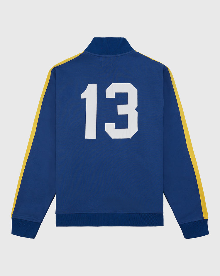 The Ebbets Field Pennsylvania Stoners Soccer Warm Up Jacket is blue with yellow sleeve stripes and a large white number 13 on the back, shown laid flat on a white background.
