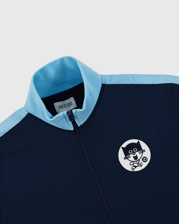 The Chicago Cats Soccer Warm Up Jacket by Ebbets Field features a navy blue design with light blue collar and shoulder stripes, and a cartoon cat face patch with bow tie and ball on the left chest. Inspired by classic American Soccer League style.