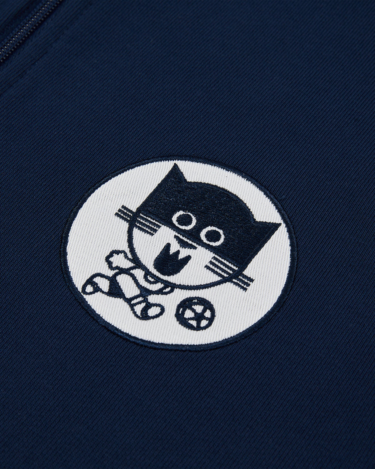 A close-up of navy blue fabric from the Ebbets Field Chicago Cats Soccer Warm Up Jacket features an embroidered white circular patch with a playful black cat holding a ball and waving.