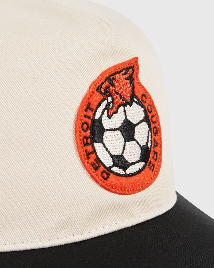 Close-up of the Detroit Cougars A-Frame Cotton Twill Ballcap by Ebbets Field, featuring a beige and black retro design with an embroidered cougar head over a soccer ball and red DETROIT COUGARS text patch.