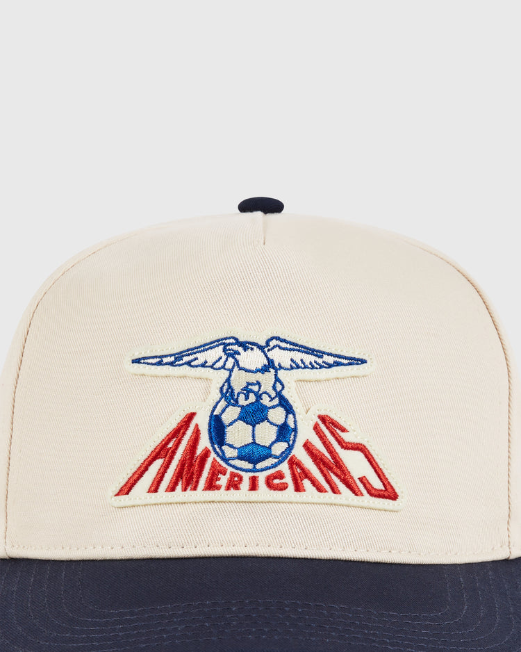 The Ebbets Field New Jersey Americans A-Frame Cotton Twill Ballcap features a beige and navy retro snapback design with an embroidered eagle on a soccer ball above bold red 