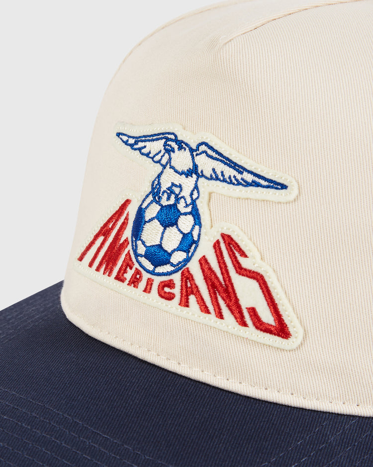 A close-up of the Ebbets Field New Jersey Americans A-Frame Cotton Twill Ballcap shows a cream cap with a dark blue brim and a patch featuring an eagle on a soccer ball above the red 