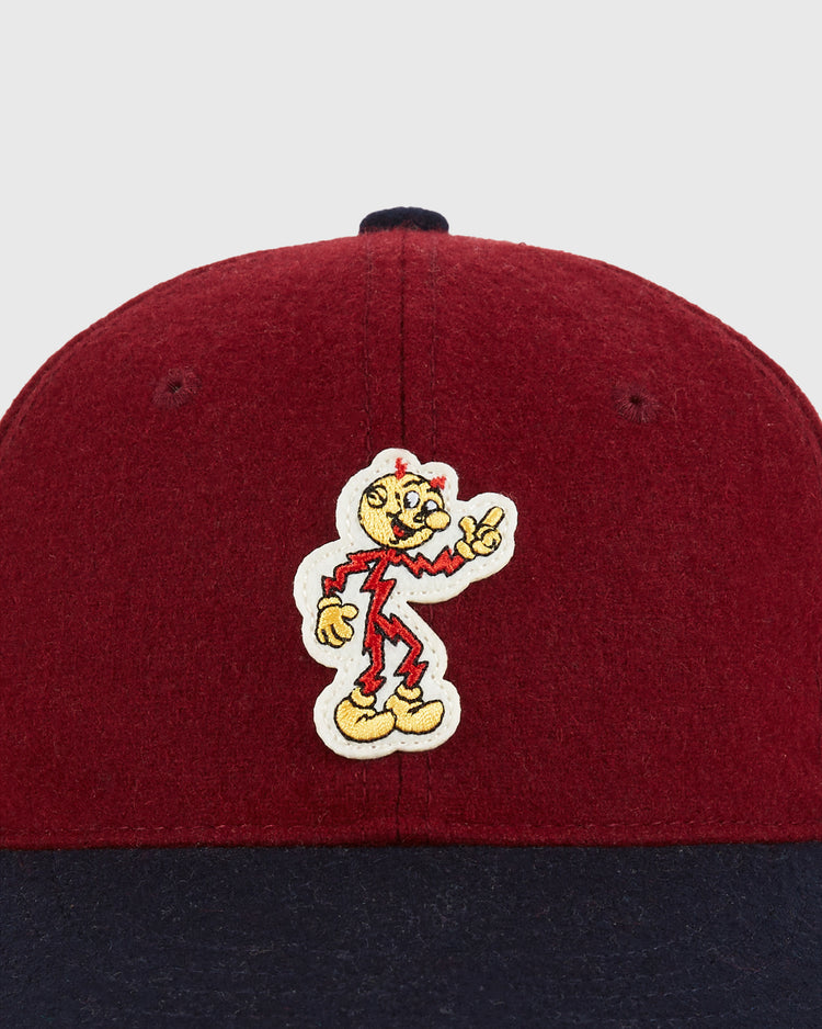 A close-up of the Ebbets Field Red Reddy K 1940 Authentic Ballcap, featuring a yellow cartoon character in a red outfit making a peace sign, embroidered on its red and dark blue cap.
