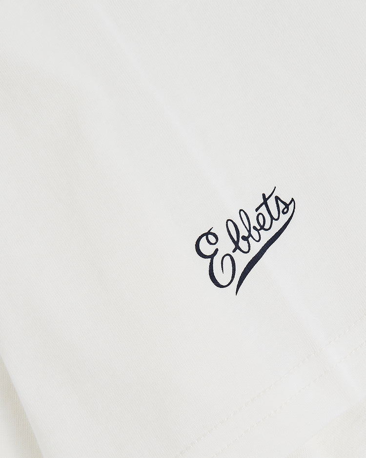 Close-up of a white fabric, likely the Sam Adams Bottle Cap Short Sleeve Graphic T-Shirt by Ebbets Field, featuring “Ebbets” in black script referencing both the brand and classic Sam Adams design.