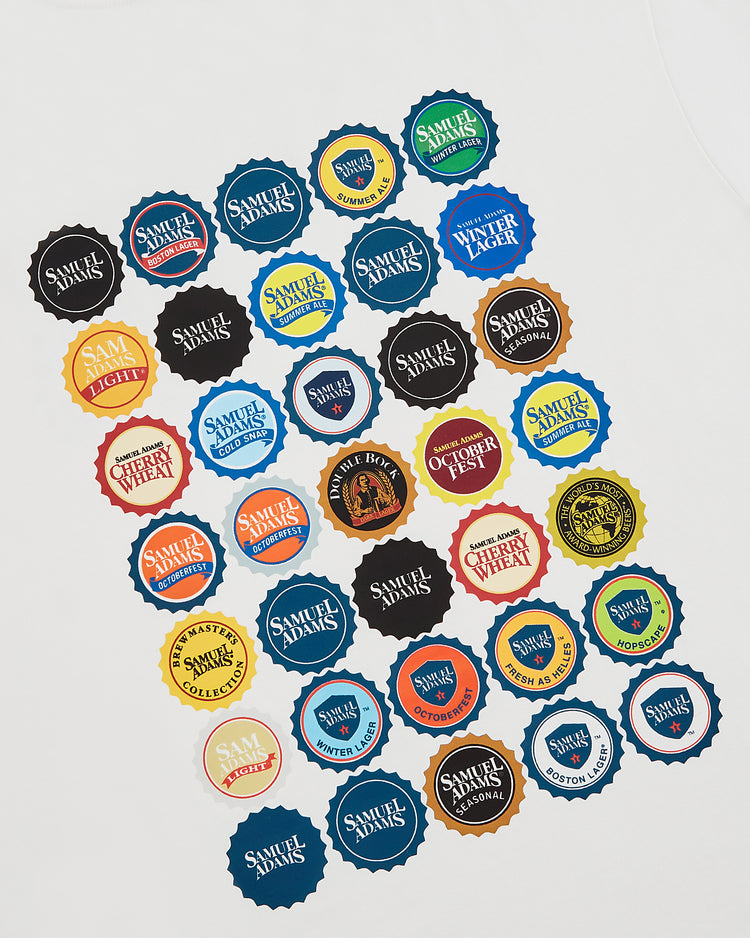 A white surface shows an assortment of Ebbets Field’s Sam Adams Bottle Cap Short Sleeve Graphic T-Shirt stickers, featuring vibrant beer label designs like Lager, Octoberfest, and Winter Lager in a scattered display.