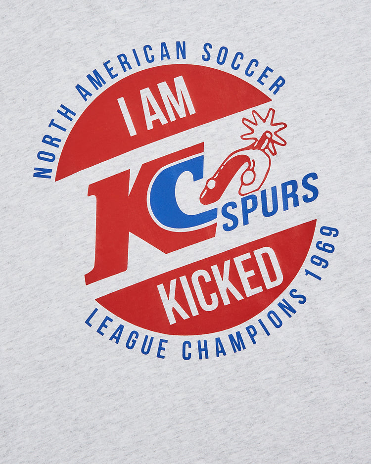 The Ebbets Field Kansas City Spurs Short Sleeve Graphic T-Shirt features a gray design with a red and blue circular logo—reading “North American Soccer League Champions 1969, I Am Kicked, KC Spurs”—and a cartoon kicking leg.