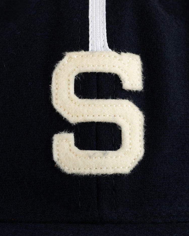 A close-up of a white stitched “S” patch with a vertical stripe on dark fabric, from the EFF Sacramento Solons 1945 Authentic Wool Softbrim Ballcap.