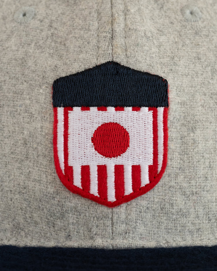 A close-up of gray fabric on the EFF U.S. Tour of Japan 1922 Authentic Wool Ballcap, featuring an embroidered shield with red stripes, a central red circle, and a navy blue top border inspired by vintage baseball memorabilia.