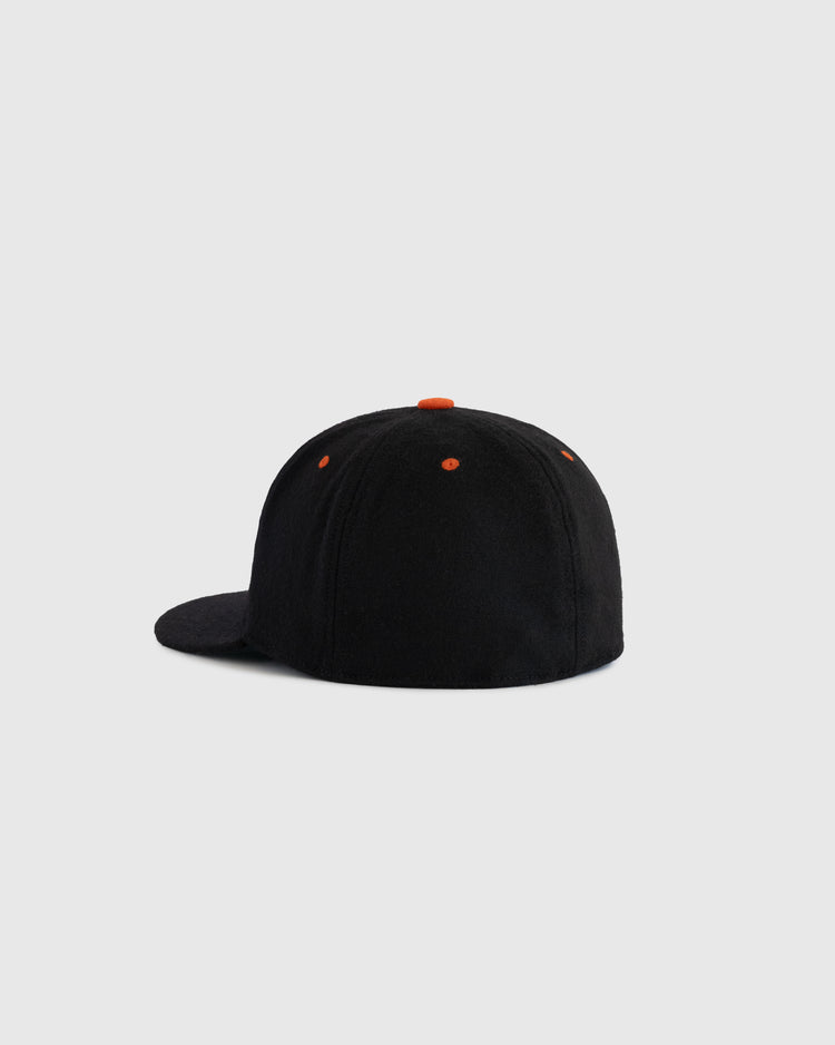 An EFF Los Angeles Aztecs 1976 Authentic Wool Ballcap in black is shown from the back, featuring orange accents on the top button and eyelets, inspired by the iconic NASL soccer team, set against a plain light gray background.