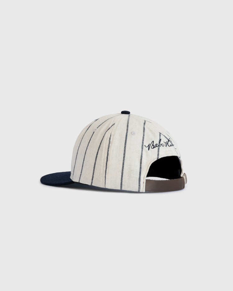 The Babe Ruth 1932 Signature Series Wool Ballcap by Ebbets Field Flannels features a cream color with thin navy pinstripes, a navy brim, brown adjustable strap, and Babe Ruths embroidered signature on the back. Displayed on a white background.