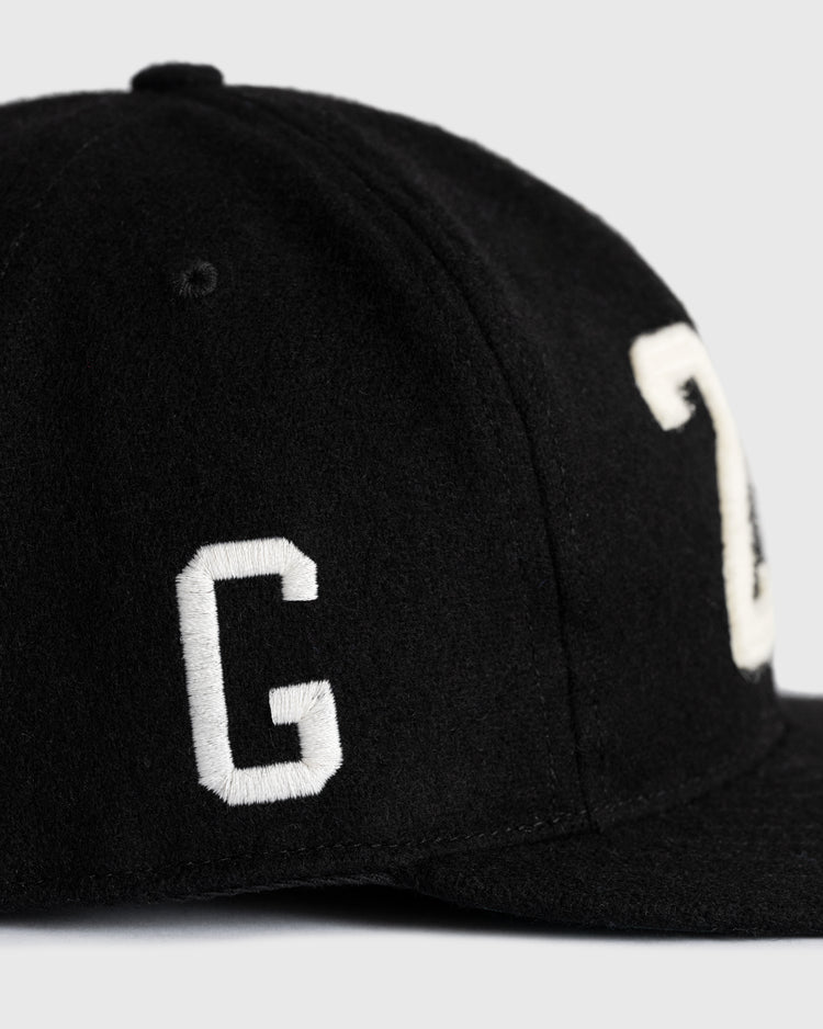 A close-up of the Ebbets Field Flannels Josh Gibson 1945 Signature Series Wool Ballcap, a black vintage-style cap with large white embroidered G and partial C, inspired by Negro Baseball Leagues, shown against a plain light background.