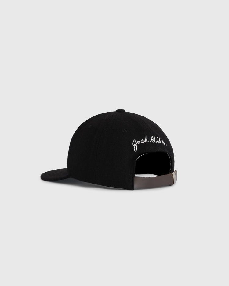 The Josh Gibson 1945 Signature Series Wool Ballcap by Ebbets Field Flannels is a black baseball cap with vintage inspiration, white cursive embroidery on the left side, and an adjustable brown strap with metal buckle. Plain light gray background.
