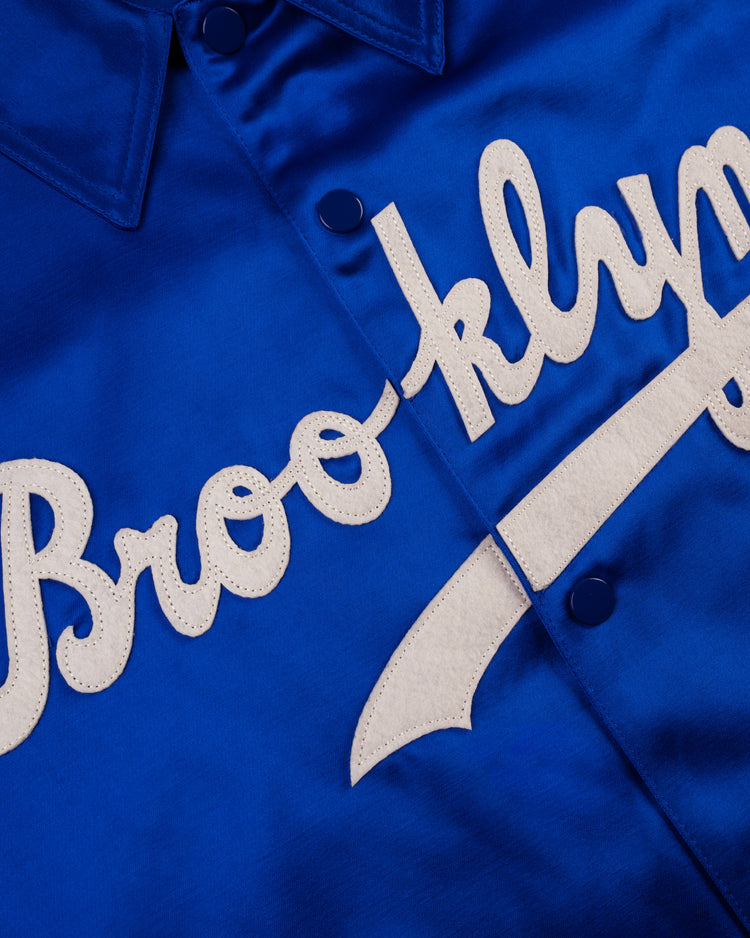 Close-up of the EFF Brooklyn Dodgers 1945 Satin Windbreaker Jacket, featuring blue satin fabric, white Brooklyn script on the front, blue snap buttons, and a pointed collar.