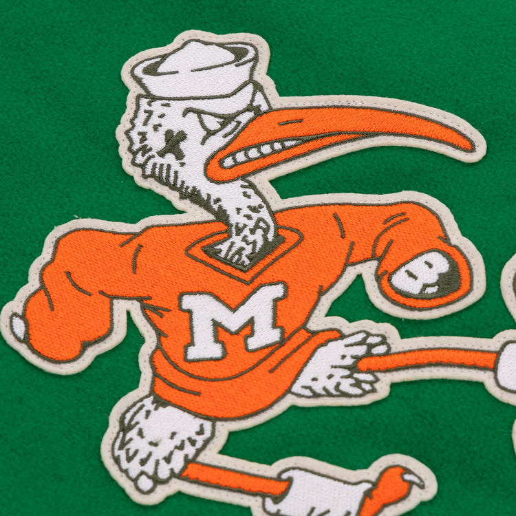 A cartoon white ibis in a sailor hat and orange sweater with a white M poses on a green background, channeling the University of Miami 1969 Authentic Jacket style by EFF—wings raised in determination.