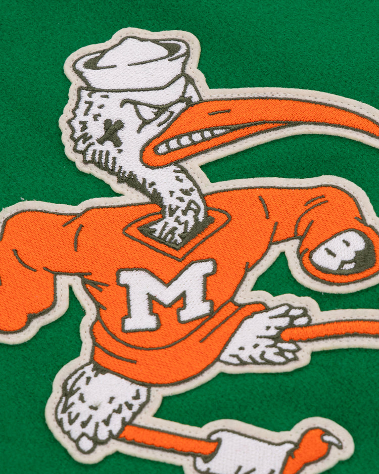 A cartoon white ibis in a sailor hat and orange sweater with a white M poses on a green background, channeling the University of Miami 1969 Authentic Jacket style by EFF—wings raised in determination.