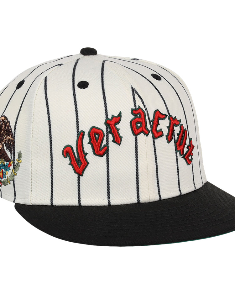 The Veracruz NLB Pinstripe Fitted Ballcap by EFF is a white cap with black pinstripes, a black brim, Veracruz in red gothic letters on the front, and an eagle-and-cactus embroidered patch on the side.