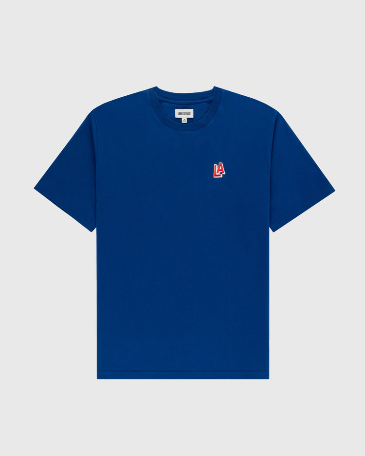 The Ebbets Field Los Angeles Angels (PCL) Embroidered ‘LA’ Short Sleeve Tee features a plain blue design with a small red and white 1942 LA team logo on the left chest, shown on a light gray background.