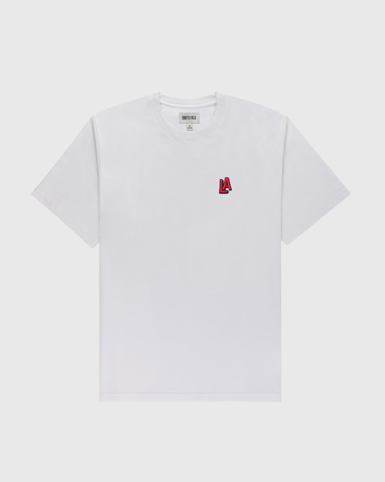 The Ebbets Field Los Angeles Angels (PCL) Embroidered ‘LA’ Short Sleeve Tee features a classic white design with a small red and black ‘LA’ logo on the left chest, laid flat on a light gray background, reflecting vintage-inspired style.