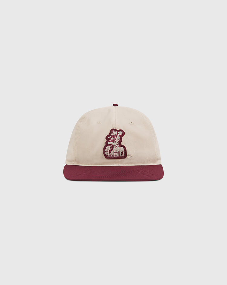 The Texas A&M Sarge Cotton Twill Ballcap by Ebbets Field is a cream-colored cap with a maroon brim, featuring an embroidered graphic of a person holding a dog on the front—an ideal choice for Texas A&M University fans looking for stylish comfort.
