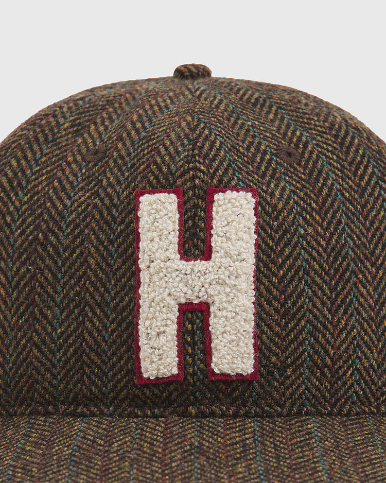 A close-up of the Harvard University Herringbone Ballcap by Ebbets Field, featuring a brown herringbone pattern and a prominent, textured white letter “H” outlined in red on the front.