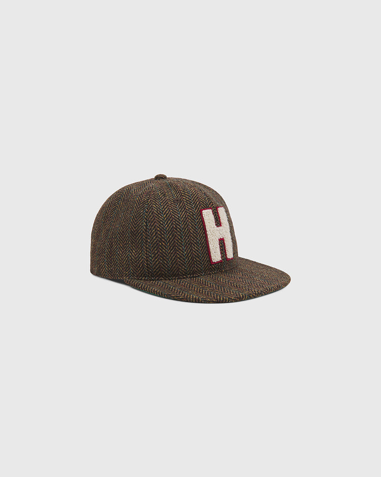 The Harvard University Herringbone Ballcap by Ebbets Field features a brown herringbone pattern, a flat brim, and a prominent white “H” outlined in red on the front. The cap is shown against a plain light gray background.