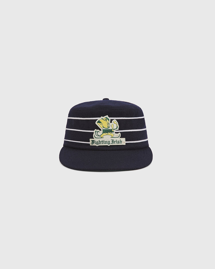 The Ebbets Field Notre Dame Pillbox Wool Ballcap is a classic navy blue hat featuring white horizontal stripes and a patch with the leprechaun mascot above the words “Fighting Irish” on the front.