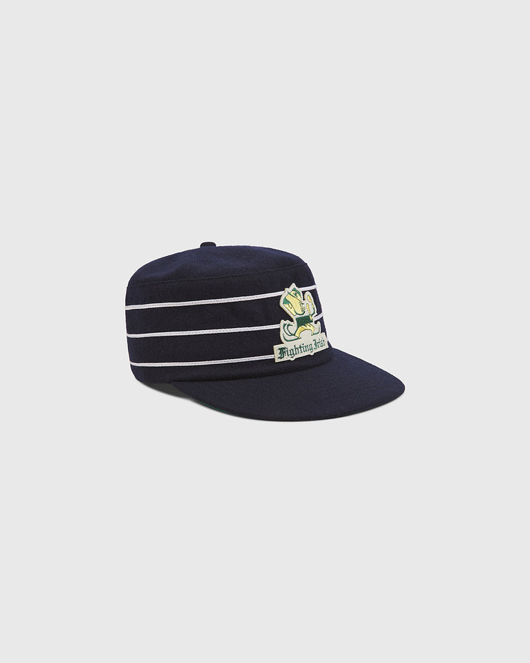 Ebbets Field’s Notre Dame Pillbox Wool Ballcap features navy blue wool with white horizontal stripes and a leprechaun mascot patch, inspired by vintage Fighting Irish pillbox hat styles. The background is plain light gray.